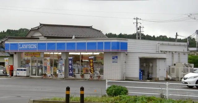 A Lawson convenience store with its blue LAWSON sign, a tiled-roof house behind it and an empty car park in front.