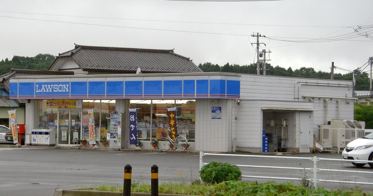 A Lawson convenience store with its blue LAWSON sign, a tiled-roof house behind it and an empty car park in front.
